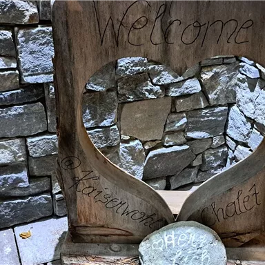 A heart-shaped welcome sign made of wood stands in front of a stone wall. It conveys a warm and inviting atmosphere.