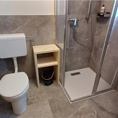 Ein modernes Badezimmer mit einer Dusche, einer Toilette und einem minimalistischen Regal. Die Wände sind mit grauen Fliesen bedeckt.
