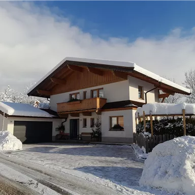 A beautiful house in the snow with a large balcony and a carport. The surroundings are wintry with plenty of snow and a blue sky.