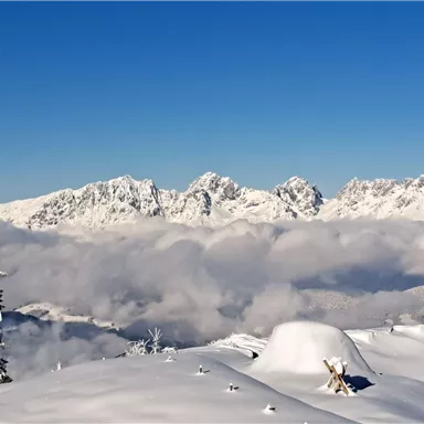 A snow-covered landscape with tall mountains and a bright blue sky. Clouds are drifting over the valleys.