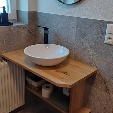 Ein modernes Badezimmer mit einem hölzernen Waschtisch und einer runden Waschschüssel. Im Hintergrund befindet sich ein runder Spiegel und eine Heizkörper.