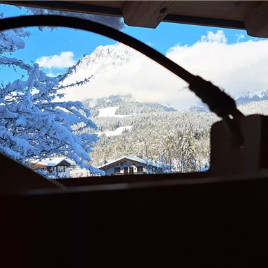 A view from a window of snow-covered mountains and a blue sky. In the foreground, a wooden element is visible.