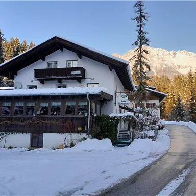 A cozy house in a winter landscape, surrounded by snow and fir trees. Majestic mountains can be seen in the background.