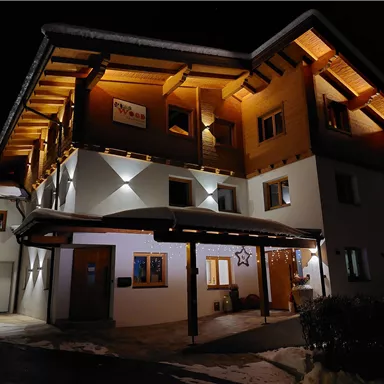A modern house with wooden details that is illuminated at night. The snow on the ground and the lights create a cozy atmosphere for the scene.