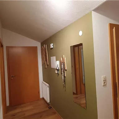 A bright hallway with a green wall and wooden floor. Pictures are hanging on the wall and there are several doors.