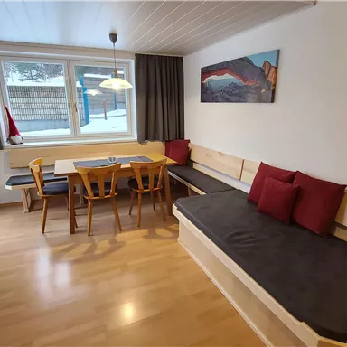 Ein gemütlicher Wohnraum mit einem Holztisch und Stühlen. Ein Sofa mit roten Kissen und große Fenster sorgen für natürliche Beleuchtung.
