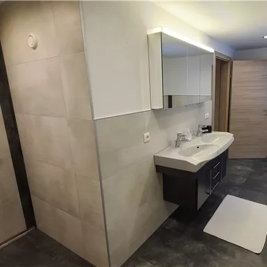 Ein modernes Badezimmer mit einer Dusche und einem großen Spiegel. Die Wände sind in hellem Steinlook und der Boden ist dunkel.