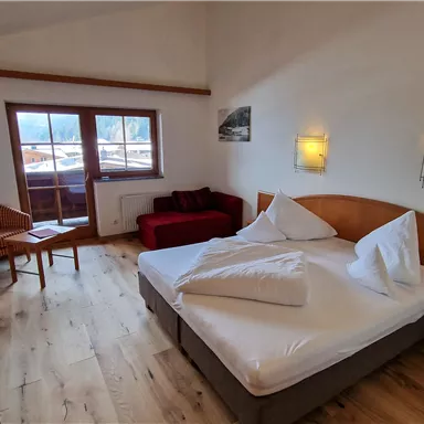 A modern hotel room with a double bed, a cozy seating area, and a flat-screen TV. Large windows provide plenty of natural light and a view outside.