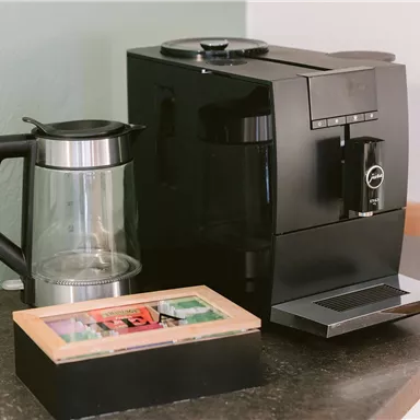 Eine schwarze Kaffeemaschine steht auf einer Küchenoberfläche neben einer Glaskanne und einer kleinen Holzbox. Die Szene ist einfach und funktional eingerichtet.