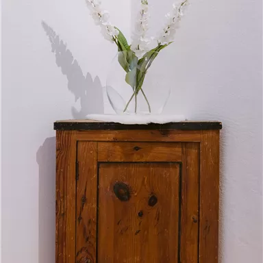 Ein rustikaler Holzschrank steht in einer Ecke. Darauf sind weiße Blumen in einer Vase platziert.