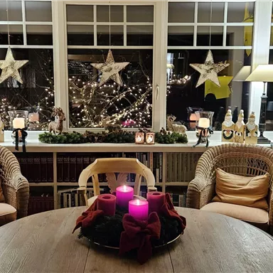 Ein gemütlicher Raum mit einem runden Tisch und zwei Korbsesseln. Der Blick aus dem Fenster zeigt festliche Dekorationen, darunter leuchtende Sterne und Kerzen.
