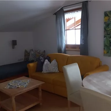 Ein modernes Wohnzimmer mit einem gelben Sofa und einem runden Esszimmer Tisch. Es gibt einen Schlafbereich und ein großes Fenster mit Vorhängen.