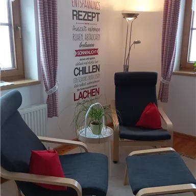 Ein gemütlicher Raum mit zwei Sesseln und einem kleinen Tisch. An der Wand befinden sich dekorative Wörter und Pflanzen.