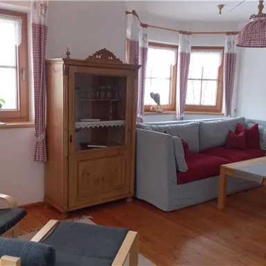 Ein gemütlicher Wohnraum mit einem Sofa in Grau und Rot sowie einem Holzkaffee Tisch. Große Fenster lassen viel Licht ein, und eine Pflanzen auf der Fensterbank fügt einen natürlichen Akzent hinzu.