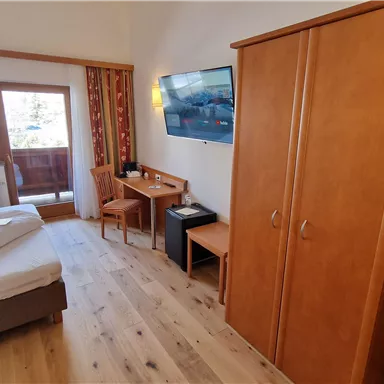 A cozy hotel room with a large bed and a desk. There is a television and a wooden wardrobe.