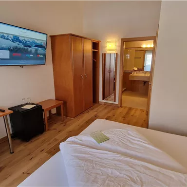 A modern hotel room with a double bed, a television, and a desk. In the background, a bathroom is visible.