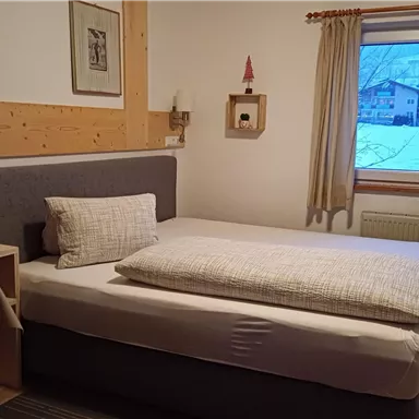 Ein einfaches Schlafzimmer mit einem Einzelbett und einem Tisch. Durch das Fenster sieht man eine verschneite Landschaft.