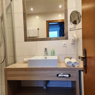 Ein modernes Badezimmer mit einem Holzwaschtisch und einem großen Spiegel. Die Dusche ist ebenfalls sichtbar, und es liegen Handtücher bereit.