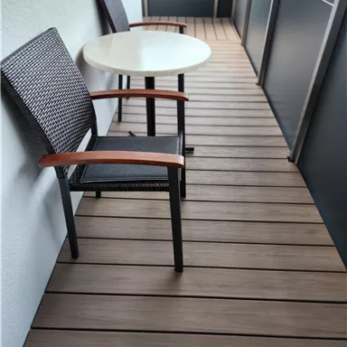 Ein Balkon mit zwei Stühlen und einem Tisch. Der Boden ist aus Holzplanken und die Wände sind teilweise abgedeckt.