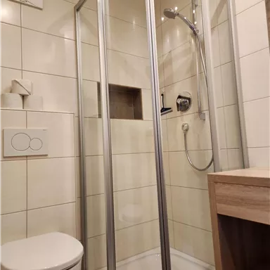 Ein modernes Badezimmer mit einer Duschkabine und einer Toilette. Die Wände sind hell gefliest und es gibt eine Holzablage.
