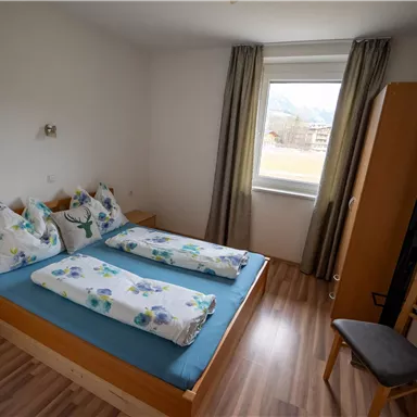 Ein einfaches Schlafzimmer mit zwei Einzelbetten und einem Fenster. Es gibt eine Kommode und einen Stuhl im Raum.