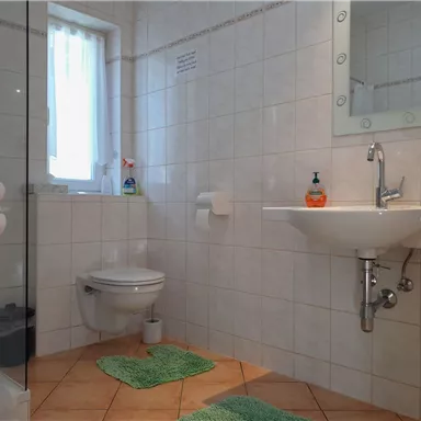 A modern bathroom with white tiles and a large mirror. There is a shower, a sink, and a toilet, all clean and well organized.