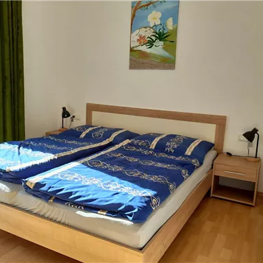 A cozy bedroom with a double bed and blue bedcovers. A painting hangs on the wall and there is a nightstand with a lamp.