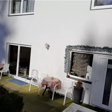 A terrace with white chairs and tables. The background shows a white wall and windows.