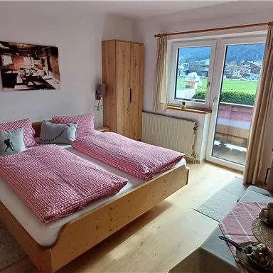 Ein gemütliches Schlafzimmer mit einem Doppelbett, hellem Holz und sanften Farben. Es gibt einen Balkon mit Blick auf die Natur und eine kleine Sitzecke.