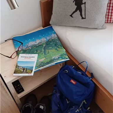 Ein gemütliches Zimmer mit einem Kissen, das einen Wanderer zeigt. Neben einem Rucksack und Wanderschuhen liegt eine Karte auf einem Nachtkästchen.