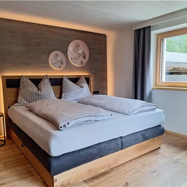 A modern bedroom with a large double bed and cozy cushions. The windows let in plenty of daylight, and the walls are decorated in neutral colors.
