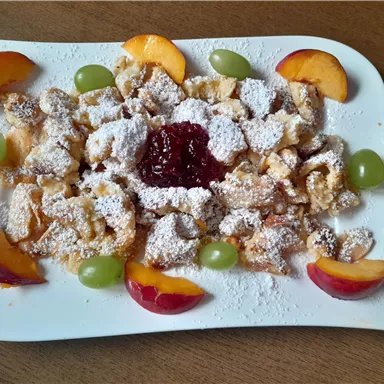 Ein köstliches Dessert mit gebackenem Teig, bestreut mit Puderzucker und serviert mit Früchten und Marmelade. Die bunte Anordnung von Pfirsichen und Trauben macht das Gericht ansprechend.