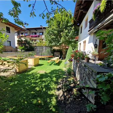 Ein schöner Garten mit grünem Gras und Bäumen. Im Hintergrund sind gemütliche Sitzgelegenheiten und typisch alpine Gebäude zu sehen.