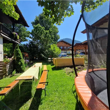 Ein schöner Garten mit Rasen, einer langen Tafel und Bänken. Im Hintergrund sieht man ein Trampolin und einige Gebäude in der Nähe.