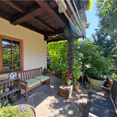 Eine gemütliche Terrasse mit Holzbank und bunten Blumen. Der Bereich ist umgeben von grünen Pflanzen und bietet eine einladende Atmosphäre.