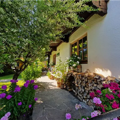 Ein gemütliches Haus mit blühenden Blumen und einem schön gestalteten Garten. Holzstapel und Bäume fügen sich harmonisch in die entspannte Atmosphäre ein.