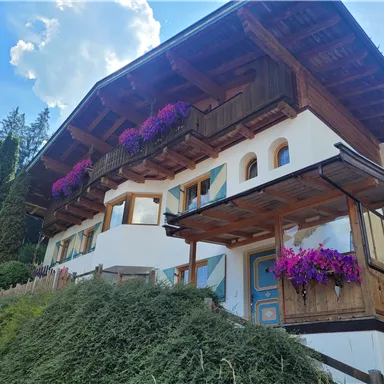 Ein traditionelles Holzhaus mit blühenden Pflanzen auf dem Balkon. Umgeben von grünen Bäumen und einem blauen Himmel.