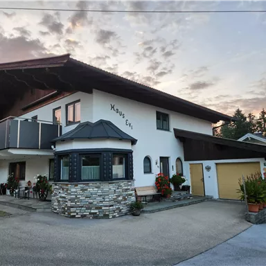 Ein großes, modernes Haus mit einem Steinsockel und Balkon. Der Himmel ist leicht bewölkt und es gibt liebevoll gestaltete Blumenbeete.