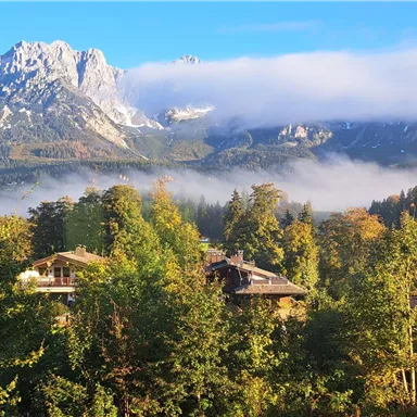 A picturesque mountain landscape with high peaks and clouds. In the foreground, green trees and some houses can be seen.