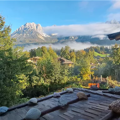 An idyllic landscape with mountains in the background and a clear blue sky. In the foreground, there are trees and a wooden building.