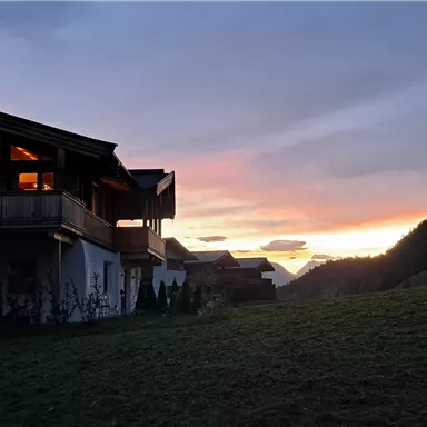 A picturesque house in the mountains during a sunset. The surroundings are green and hilly, with gentle shadows of the mountains in the background.