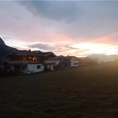 A peaceful landscape with mountains in the background and a sunset. In the foreground, houses can be seen on a meadow.