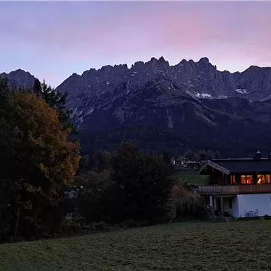 A picturesque sunset over majestic mountains. In the foreground stands a cozy house, surrounded by trees and meadows.