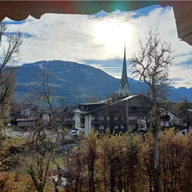 Ein malerischer Blick auf Berge und einen historischen Kirchturm. Die Sonne scheint durch die Wolken und verleiht der Landschaft eine gemütliche Atmosphäre.