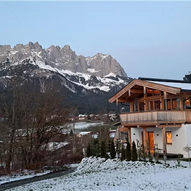 A tranquil alpine landscape with snow-capped mountains in the background. In the foreground stands a modern house with warm light and a beautiful balcony.