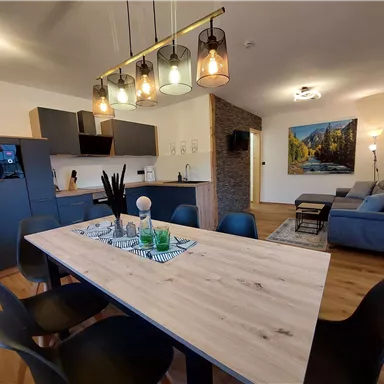 Modernes Wohnzimmer mit offener Küche und Essbereich. Helle Farben, gemütliche Möbel und eine kunstvolle Wanddekoration schaffen eine einladende Atmosphäre.