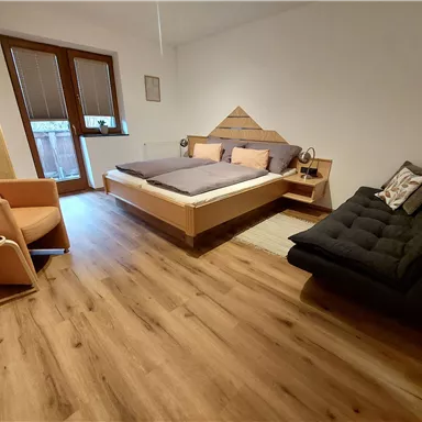 Ein modernes Schlafzimmer mit einem Doppelbett und einem Sofa. Der Raum ist hell und verfügt über große Fenster und einen warmen Holzfußboden.