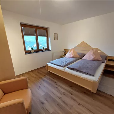Ein gemütliches Schlafzimmer mit einem Doppelbett und zwei Kissen in sanften Farben. Ein orangefarbener Sessel und ein Fenster mit Aussicht vervollständigen den Raum.