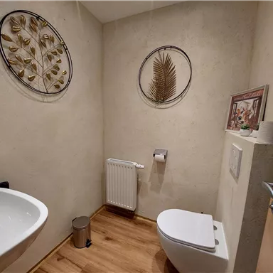 Ein modernes Badezimmer mit einer Toilette und einem Waschbecken. An den Wänden hängen dekorative Bilder und es gibt einen Holzfußboden.