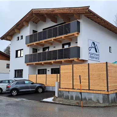 Ein modernes Gebäude mit mehreren Balkonen und einer Holzverkleidung. Vor dem Gebäude stehen einige Autos geparkt.